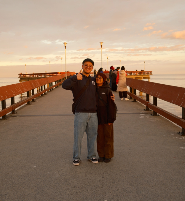 Regina and Andrew standing together on a wooden pier at sunset, Andrew giving a thumbs up