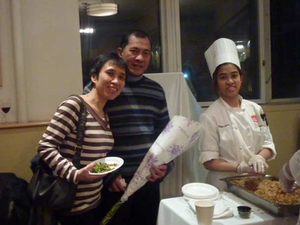 Regina in chef's whites and toque behind a banquet table, standing with her parents on the final day of her Culinary Arts program